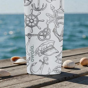 May include: White water bottle with a nautical-themed pattern of anchors, ship wheels, and rope knots. The bottle has the word "owala." printed on it. The background features a wooden dock, seashells, and the ocean.