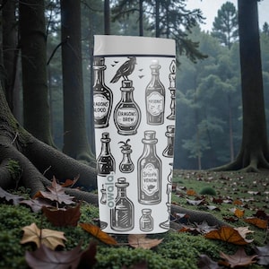 K&ouml;nnte beinhalten: Eine wei&szlig;e Owala-Trinkflasche mit einem sich wiederholenden Muster aus Zaubertr&auml;nken, einem Raben und Kerzen. Die Flaschen sind mit Namen wie "Drachenblut" und "Spinnengift" beschriftet. Die Flasche befindet sich vor einem Waldhintergrund.