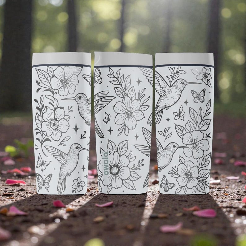 Puede incluir: Tres botellas de agua blancas Owala con dibujos lineales negros de colibr&iacute;es, flores y mariposas. Las botellas tienen una tapa blanca y un borde negro. Est&aacute;n sobre una superficie marr&oacute;n con p&eacute;talos de flores rosas.