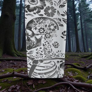 May include: A white tumbler featuring a detailed black and gray floral skull design. The design includes flowers, leaves, and a rib cage, creating a gothic and botanical theme. The tumbler is set against a blurred forest background.