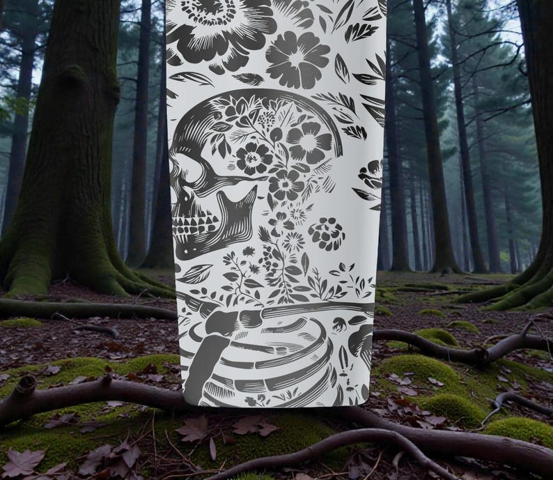 May include: A white tumbler featuring a detailed black and gray floral skull design. The design includes flowers, leaves, and a rib cage, creating a gothic and botanical theme. The tumbler is set against a blurred forest background.