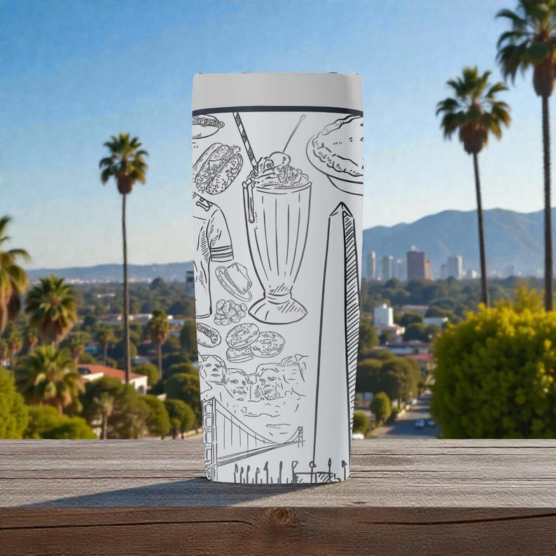 May include: A white travel mug with a black and white line drawing of American icons, including a milkshake, hot dog, and the Washington Monument. The background shows a scenic view of palm trees and a city.