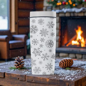 Puede incluir: Un termo blanco con tapa, decorado con un patr&oacute;n de copos de nieve negros. El termo cil&iacute;ndrico est&aacute; sobre una superficie de madera. El fondo incluye una chimenea y pi&ntilde;as, sugiriendo un tema invernal.