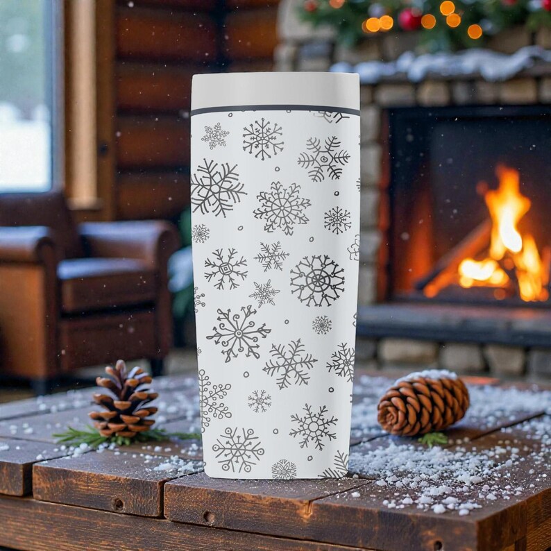 Puede incluir: Un termo blanco con tapa, decorado con un patr&oacute;n de copos de nieve negros. El termo cil&iacute;ndrico est&aacute; sobre una superficie de madera. El fondo incluye una chimenea y pi&ntilde;as, sugiriendo un tema invernal.