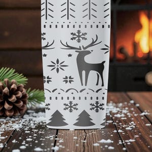 May include: A white tumbler with a black winter-themed design. The design includes a reindeer, snowflakes, and trees. The tumbler is cylindrical and appears to be made of metal. The background includes a fireplace, pine needles, and a pine cone.