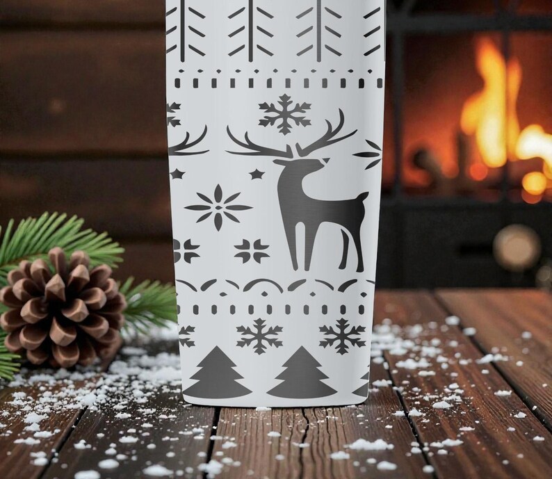 May include: A white tumbler with a black winter-themed design. The design includes a reindeer, snowflakes, and trees. The tumbler is cylindrical and appears to be made of metal. The background includes a fireplace, pine needles, and a pine cone.