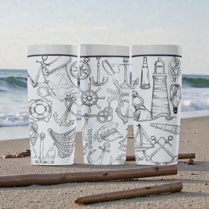 May include: Three white travel mugs with a nautical theme, featuring black line drawings of anchors, compasses, lighthouses, and sailboats. The mugs have a white lid and the brand name "owala" is visible on one of the mugs. The mugs are on a sandy beach.