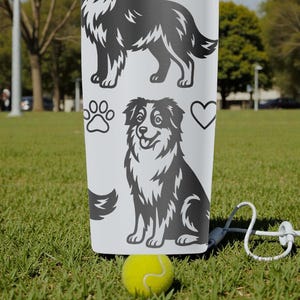 Puede incluir: Un vaso blanco con ilustraciones en blanco y negro de un perro pastor australiano, huellas de patas y un coraz&oacute;n. Una pelota de tenis amarilla y una correa blanca est&aacute;n en primer plano, sobre un campo de hierba.