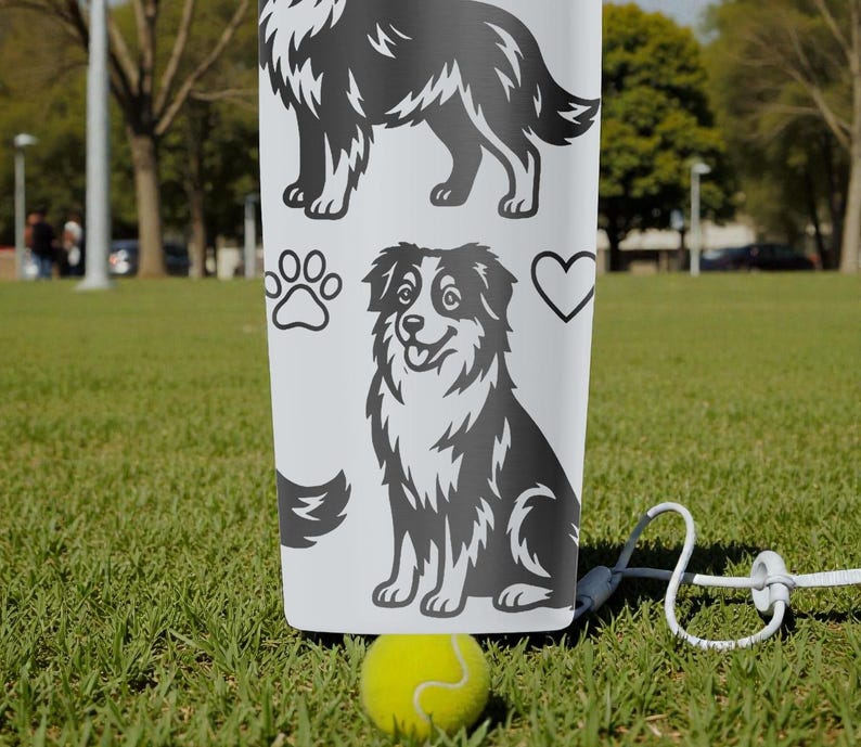 Puede incluir: Un vaso blanco con ilustraciones en blanco y negro de un perro pastor australiano, huellas de patas y un coraz&oacute;n. Una pelota de tenis amarilla y una correa blanca est&aacute;n en primer plano, sobre un campo de hierba.
