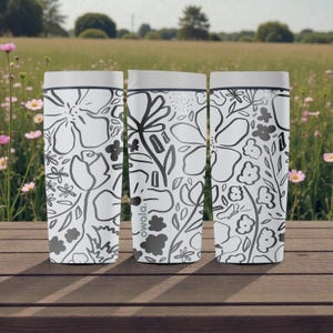 May include: Three white insulated travel mugs with a black and grey floral design. The mugs have grey lids and are on a wooden surface, with a field and flowers in the background. The brand name 'owala' is visible.
