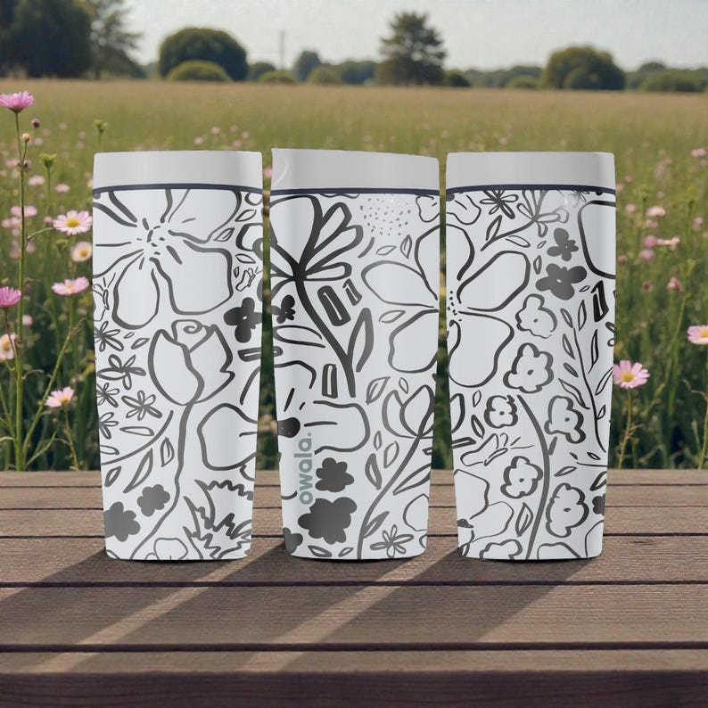 May include: Three white insulated travel mugs with a black and grey floral design. The mugs have grey lids and are on a wooden surface, with a field and flowers in the background. The brand name 'owala' is visible.