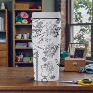 May include: White travel mug featuring a black and white illustration of sewing tools and floral designs. The design includes scissors, spools of thread, a measuring tape, and a dress form. The mug is on a wooden table.
