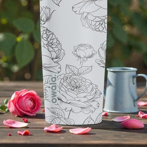 Op de afbeelding: Een witte Owala waterfles met een zwart bloemenontwerp, naast een roze roos en een blauwe mok. De fles heeft een herhalend patroon van rozen en bladeren. De sc&egrave;ne speelt zich af op een houten oppervlak met verspreide roze bloemblaadjes.