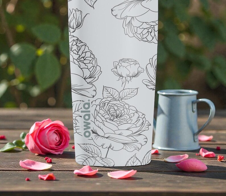 Op de afbeelding: Een witte Owala waterfles met een zwart bloemenontwerp, naast een roze roos en een blauwe mok. De fles heeft een herhalend patroon van rozen en bladeren. De sc&egrave;ne speelt zich af op een houten oppervlak met verspreide roze bloemblaadjes.
