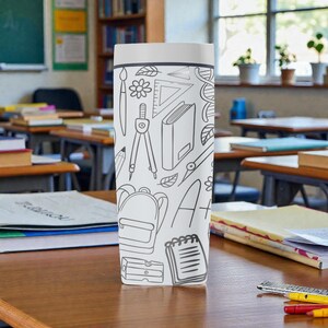 Op de afbeelding: Een wit en grijze reismok met een schoolthema-ontwerp. Het ontwerp bevat illustraties van een rugzak, passer, liniaal, potlood en andere schoolbenodigdheden. De mok is cilindrisch met een wit deksel.