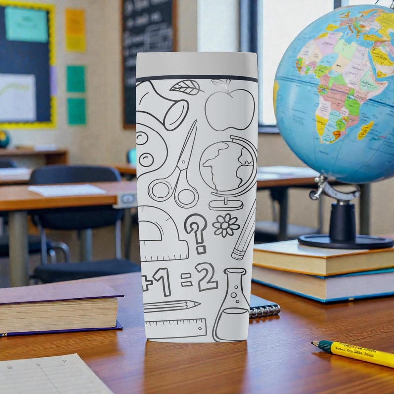 Op de afbeelding: Een witte beker met een grijze deksel, versierd met zwarte lijntekeningen van schoolgerelateerde items zoals een appel, schaar, een wereldbol en een potlood. De beker staat op een houten bureau in een klaslokaal.
