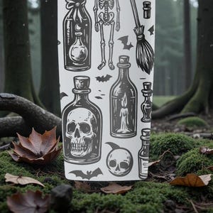 K&ouml;nnte beinhalten: Ein wei&szlig;er Becher mit einem schwarz-grauen Halloween-Design. Das Design umfasst Illustrationen von Zaubertr&auml;nken, einem Skelett, Flederm&auml;usen, einem Besen und einem Totenkopf. Der Becher steht vor einem Hintergrund aus Waldboden mit Moos und Herbstlaub.