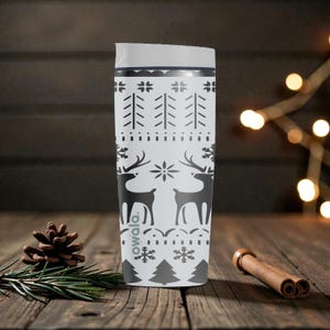 May include: A white travel mug with a black and gray winter pattern featuring reindeer, trees, and snowflakes. The mug has a white lid and a silver band near the top. The brand name "owala" is printed on the side.
