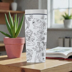 May include: White travel mug featuring black line art of characters and elements from the animated film "Tangled." The design includes Rapunzel, Pascal the chameleon, and floating lanterns. The mug has a white lid and a rectangular shape.