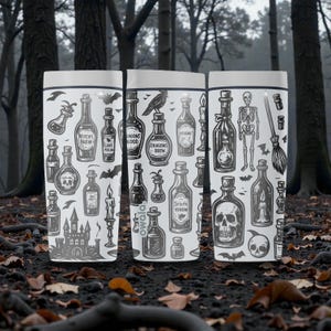 K&ouml;nnte beinhalten: Drei wei&szlig;e Becher mit einem gruseligen Halloween-Thema. Jeder Becher zeigt ein anderes Design von Zaubertr&auml;nken, Totenk&ouml;pfen, Flederm&auml;usen und einem Skelett. Die Becher stehen vor einem verschwommenen Waldboden.
