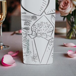 May include: A white bottle with black line art illustrations of a bouquet, microphone, and key. The word "owala" is printed on the bottle. Pink rose petals are scattered on the table, with a champagne glass in the background.