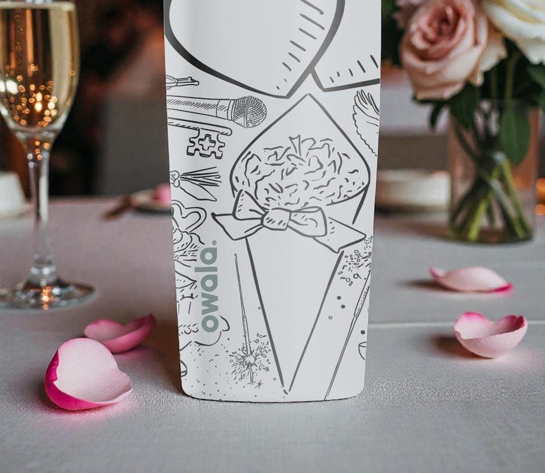 May include: A white bottle with black line art illustrations of a bouquet, microphone, and key. The word "owala" is printed on the bottle. Pink rose petals are scattered on the table, with a champagne glass in the background.
