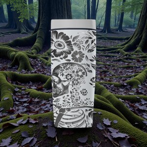May include: A white travel mug with a black floral and skull design. The design includes large flowers, leaves, and a skull filled with floral patterns. The bottom of the mug features a rib cage design. The mug has a white lid and a black rim.