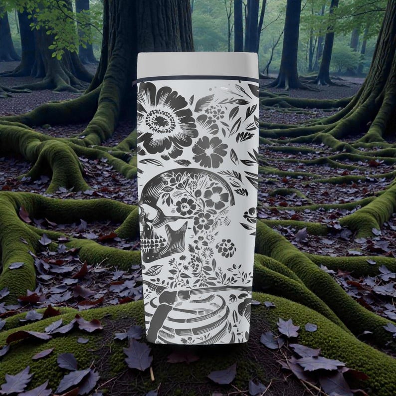 May include: A white travel mug with a black floral and skull design. The design includes large flowers, leaves, and a skull filled with floral patterns. The bottom of the mug features a rib cage design. The mug has a white lid and a black rim.