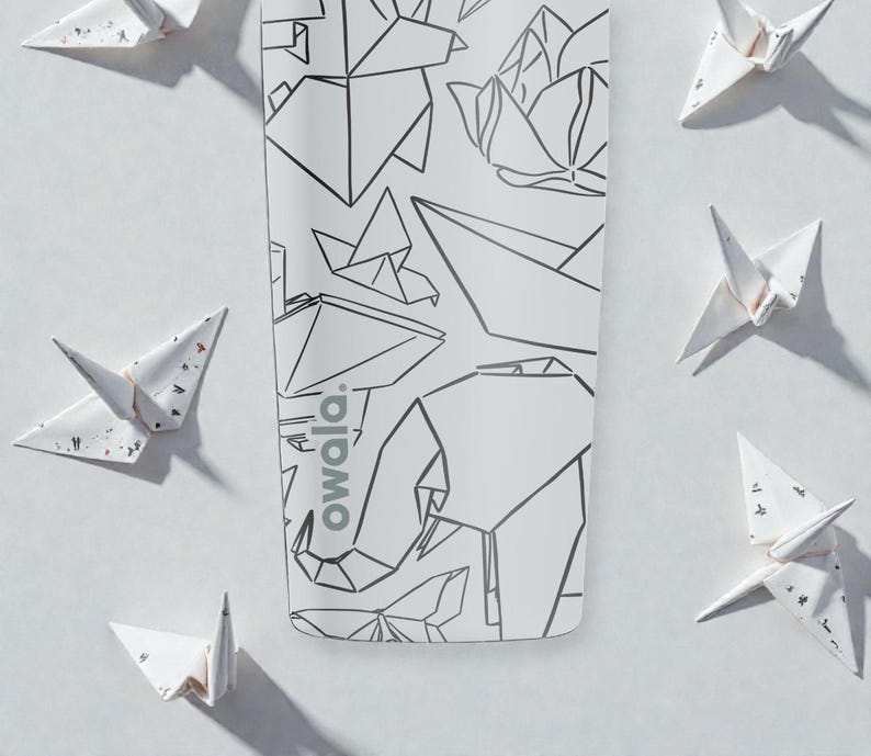 K&ouml;nnte beinhalten: Wei&szlig;e Wasserflasche mit geometrischem Design mit einem Elefanten und anderen Formen. Auf der Flasche steht das Wort "owala". Origami-Papierkraniche sind auf wei&szlig;em Hintergrund um die Flasche herum verstreut.