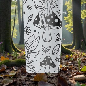 May include: A white tumbler with a black fairy and mushroom design. The design includes a fairy on a mushroom, plus other botanical elements. The tumbler is set against a blurred forest background.