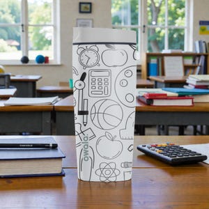 Op de afbeelding: Een witte Owala waterfles met een grijze deksel, versierd met zwarte lijntekeningen van schoolgerelateerde items zoals een wekker, appel, rekenmachine en basketbal. De fles staat op een houten bureau in een klaslokaal.