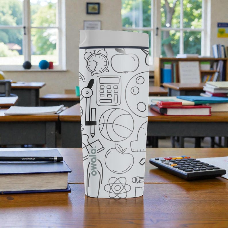 Op de afbeelding: Een witte Owala waterfles met een grijze deksel, versierd met zwarte lijntekeningen van schoolgerelateerde items zoals een wekker, appel, rekenmachine en basketbal. De fles staat op een houten bureau in een klaslokaal.