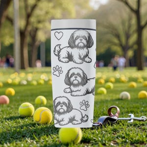 May include: A white tumbler featuring a repeating pattern of a Shih Tzu dog, paw prints, and a heart. The tumbler is cylindrical and stands upright on a grassy surface scattered with tennis balls. A dog leash and clip are visible.