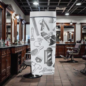 May include: A white, cylindrical banner featuring a repeating pattern of barber shop tools in black and gray. The design includes combs, razors, barber chairs, hair dryers, and scissors. The banner is displayed in a barber shop setting.