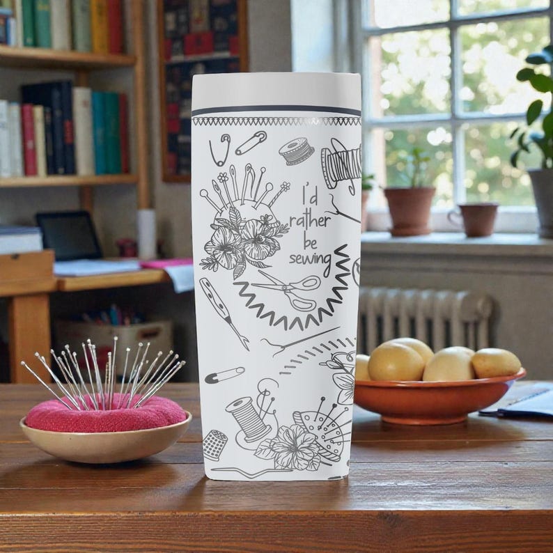May include: A white travel mug with a black and white sewing-themed design, including scissors, pins, and the text "I'd rather be sewing." A red pincushion with pins is next to the mug. The mug is on a wooden table.