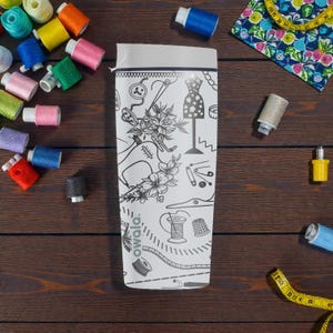 May include: A white Owala water bottle with a black and white sewing-themed design, featuring illustrations of a sewing machine, dress form, and other sewing notions. The bottle is surrounded by colorful spools of thread, a measuring tape, and fabric.