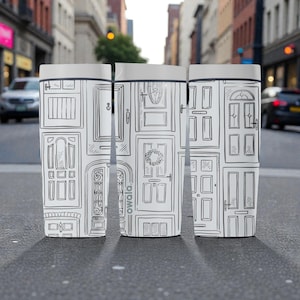 Peut inclure: Trois gobelets de voyage blancs avec un dessin au trait noir de diverses portes. Les gobelets ont un couvercle et une base gris. Les gobelets sont posés sur une surface grise avec une rue de la ville en arrière-plan.