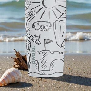 Op de afbeelding: Witte beker met zwarte lijntekeningen van strandthema's, waaronder een zon, snorkel en zandkasteel. Het woord "owala." is verticaal afgedrukt. Een zeeschelp en drijfhout liggen op het zandstrand op de voorgrond.