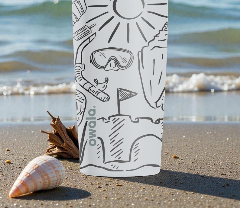 Op de afbeelding: Witte beker met zwarte lijntekeningen van strandthema's, waaronder een zon, snorkel en zandkasteel. Het woord "owala." is verticaal afgedrukt. Een zeeschelp en drijfhout liggen op het zandstrand op de voorgrond.