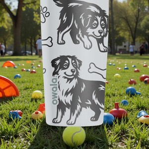Puede incluir: Una botella de agua blanca Owala con ilustraciones de perros en blanco y negro, huellas de patas y un hueso. La botella est&aacute; en un parque con una pelota de tenis y juguetes coloridos.