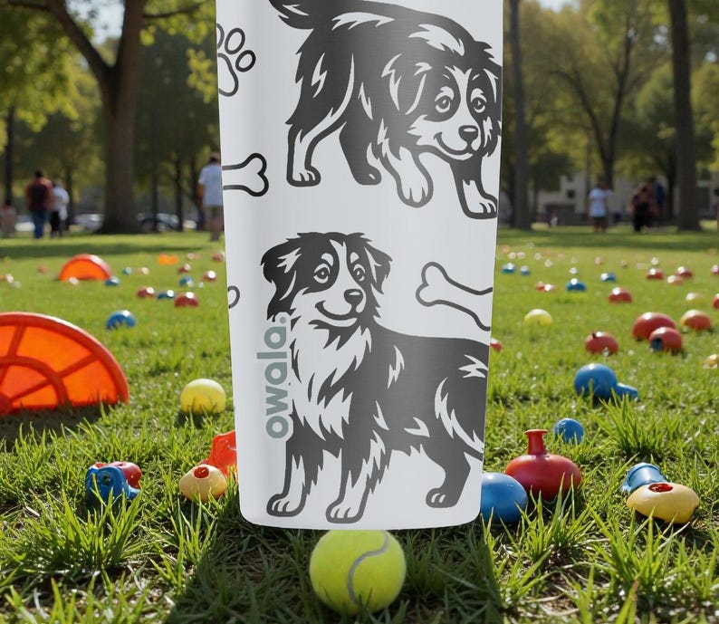 Puede incluir: Una botella de agua blanca Owala con ilustraciones de perros en blanco y negro, huellas de patas y un hueso. La botella est&aacute; en un parque con una pelota de tenis y juguetes coloridos.