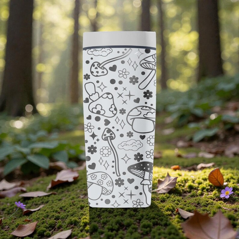 Peut inclure: Un mug de voyage blanc avec un motif de champignons noir et blanc. Le motif comprend diverses illustrations de champignons, des c&oelig;urs, des fleurs et des &eacute;toiles. Le mug est cylindrique avec un couvercle blanc, sur un fond de for&ecirc;t flou.