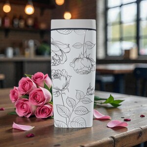 Op de afbeelding: Een witte reismok met een bloemenlijnkunstontwerp van rozen en bladeren. De mok heeft een lichtgrijze deksel en staat op een houten tafel naast een boeket roze rozen. Rozenblaadjes liggen verspreid op de tafel.