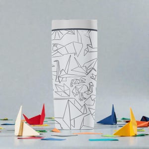 K&ouml;nnte beinhalten: Wei&szlig;er Reisebecher mit Deckel, verziert mit einem sich wiederholenden Muster aus grauen Origami-Tieren. Origami-Papierboote und bunte Papierschnipsel sind um die Basis des Bechers verstreut. Der Becher hat einen wei&szlig;en Deckel und einen grauen Rand.
