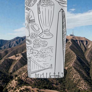 May include: A tall, white tumbler with black line drawings of American landmarks and food items, including a milkshake, donuts, and the Washington Monument. The background shows a mountain range under a blue sky.