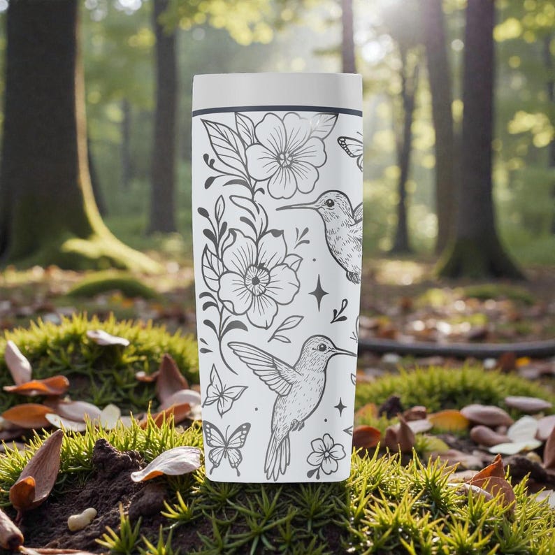 Puede incluir: Taza de viaje blanca con un dise&ntilde;o floral y colibr&iacute; en blanco y negro. El dise&ntilde;o incluye flores, mariposas y colibr&iacute;es. La taza es cil&iacute;ndrica y tiene una tapa blanca.