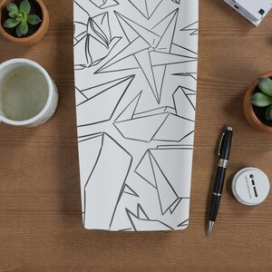 K&ouml;nnte beinhalten: Ein wei&szlig;es rechteckiges Objekt mit einem schwarzen geometrischen Design, m&ouml;glicherweise ein Notizbuch oder ein dekoratives St&uuml;ck, liegt auf einer Holzoberfl&auml;che. Eine wei&szlig;e Tasse, ein Stift und kleine Topfpflanzen sind ebenfalls sichtbar. Das Design weist eckige Formen auf.