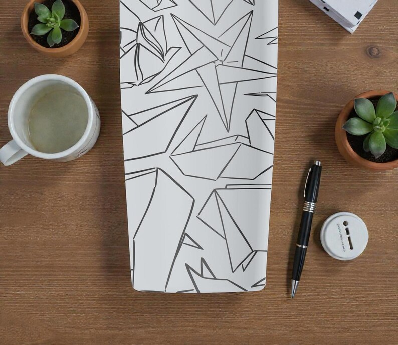 K&ouml;nnte beinhalten: Ein wei&szlig;es rechteckiges Objekt mit einem schwarzen geometrischen Design, m&ouml;glicherweise ein Notizbuch oder ein dekoratives St&uuml;ck, liegt auf einer Holzoberfl&auml;che. Eine wei&szlig;e Tasse, ein Stift und kleine Topfpflanzen sind ebenfalls sichtbar. Das Design weist eckige Formen auf.