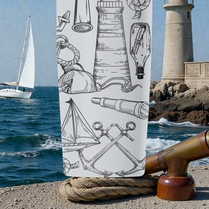 May include: A white tumbler featuring nautical illustrations of anchors, lighthouses, and sailboats. A vintage-style telescope sits on a wooden base. A sailboat is in the background, sailing on the blue ocean. The tumbler is resting on a thick rope.