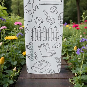 Puede incluir: Una botella de agua Owala blanca con un dise&ntilde;o de garabatos con tem&aacute;tica de jard&iacute;n. Las ilustraciones incluyen una valla, botas, una paleta y una seta. La botella est&aacute; sobre una superficie de madera, rodeada de flores.